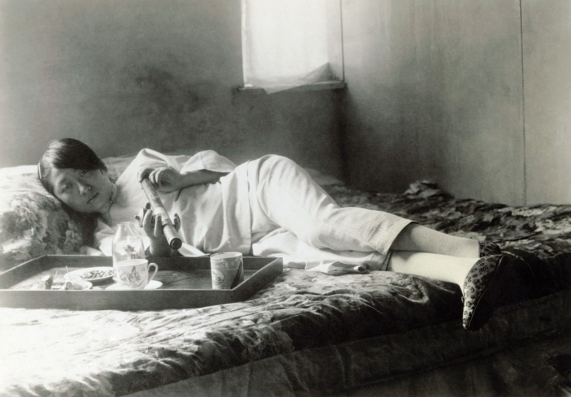 Foto einer Frau mit einer großen Pfeife, die auf einem Bett liegt und in die Kamera blickt. Das Foto wurde in China im 19. Jahrhundert aufgenommen.