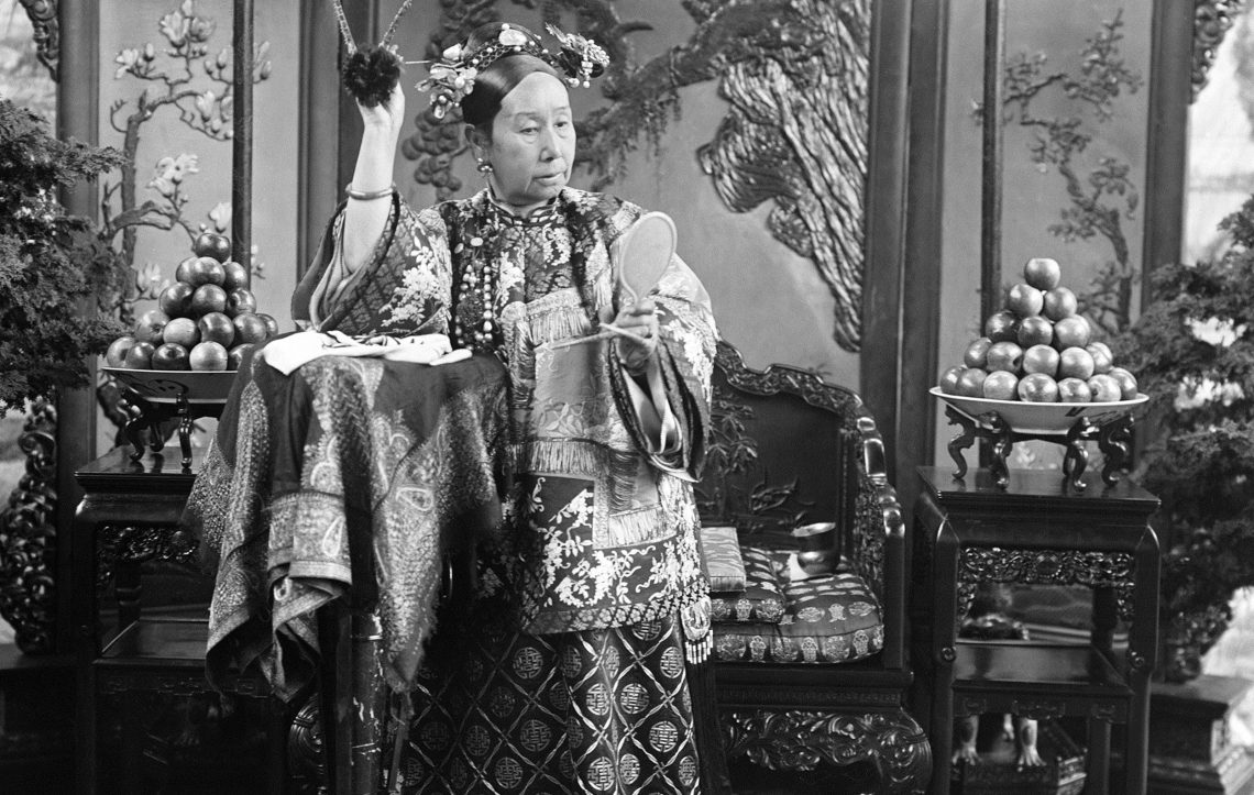 Eine Frau in aufwändiger Kleidung hält einen Handspiegel und eine Blume. Sie steht in einem prächtig eingerichteten Raum. Das Bild gehört zu einem Beitrag über Kaiserin Cixi.