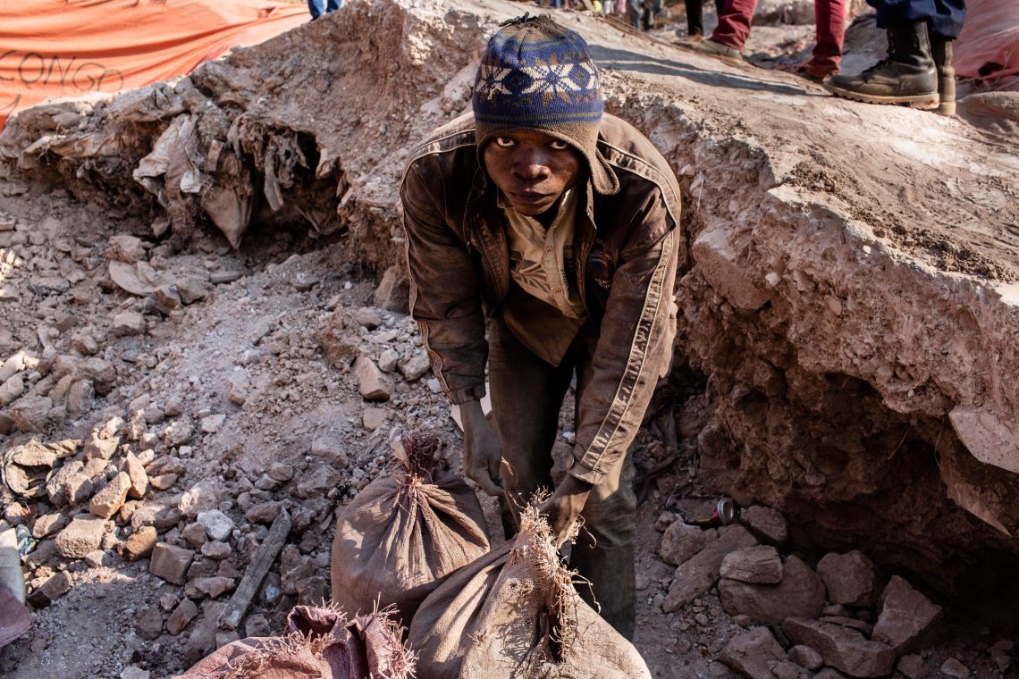 Foto eines Arbeiters in einer Mine vor Steinschutt mit gefüllten Säcken, der in die Kamera blickt. Das Bild ist Teil von einem Beitrag über die Rohstoffe, die für Elektroautos gebraucht werden.