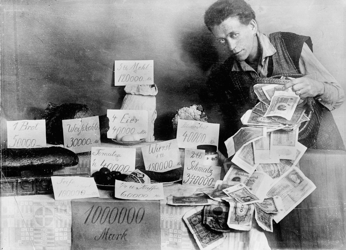 Hyperinflation während der Weimarer Republik: Eine Person schüttet kübelweise Markscheine neben einen Tisch auf dem Lebensmittel mit horrenden Preisien angeschrieben stehen.