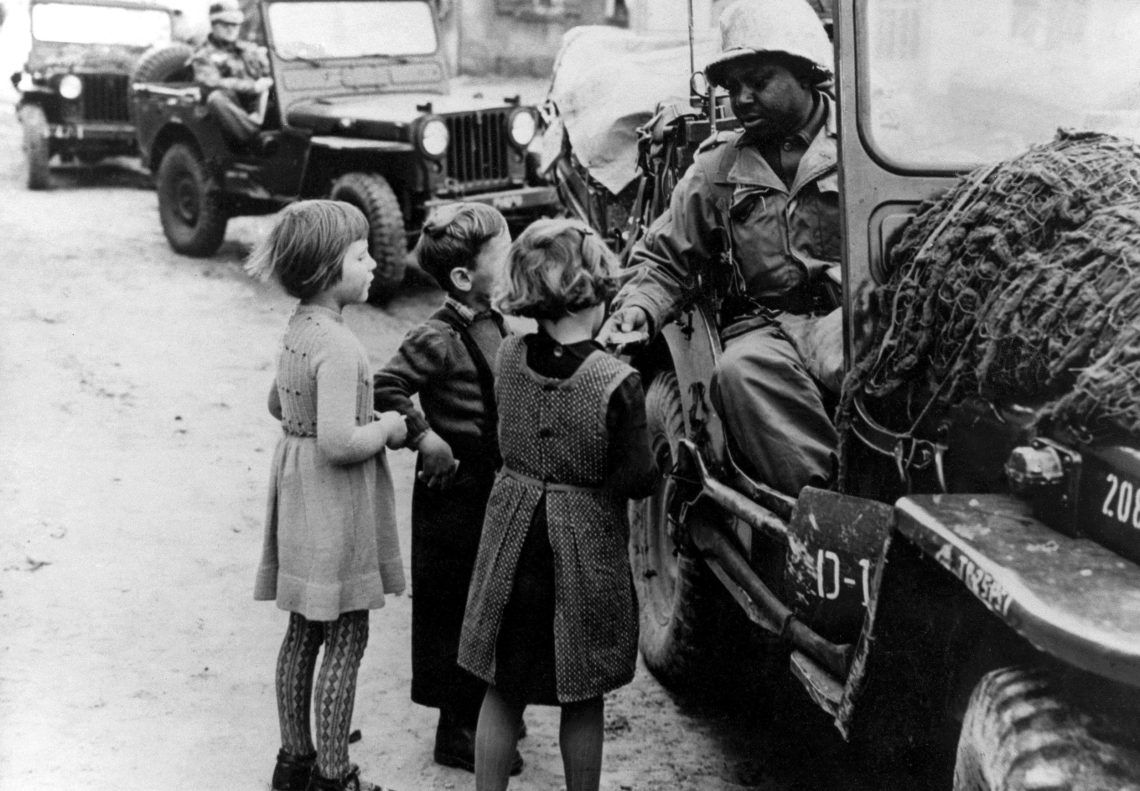 Ein Soldat in einem offenen Jeep verteilt etwas an drei Kinder, die bei seinem Jeep stehen. Im Hintergrund sind weitere Militärfahrzeuge zu sehen. Das Bild illustriert einen Beitrag über eine mögliche gemeinsame Armee in der EU.