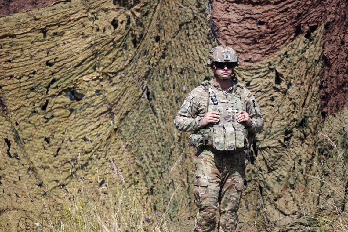Ein Soldat in Tarnmontur steht vor einem Tarnvorhang. Er trägt eine Sonnenbrille und einen Helm. Das Bild ist Teil eines Artikels über eine EU-Armee.