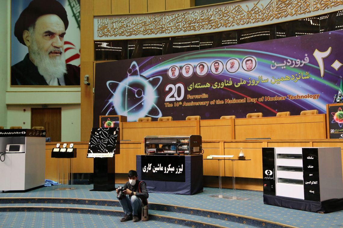 Ein Mann mit einer Kamera sitzt auf den Stufen einer Art Bühne in einem großen Raum. Im Hintergrund ist ein großes Plakat mit einem Atommodell zu sehen sowie einige Ausstellungstische. An der Wand hängt ein sehr großes Plakat von Ajatollah Khomeini. Das Bild gehört zu einem Beitrag über den Iran.