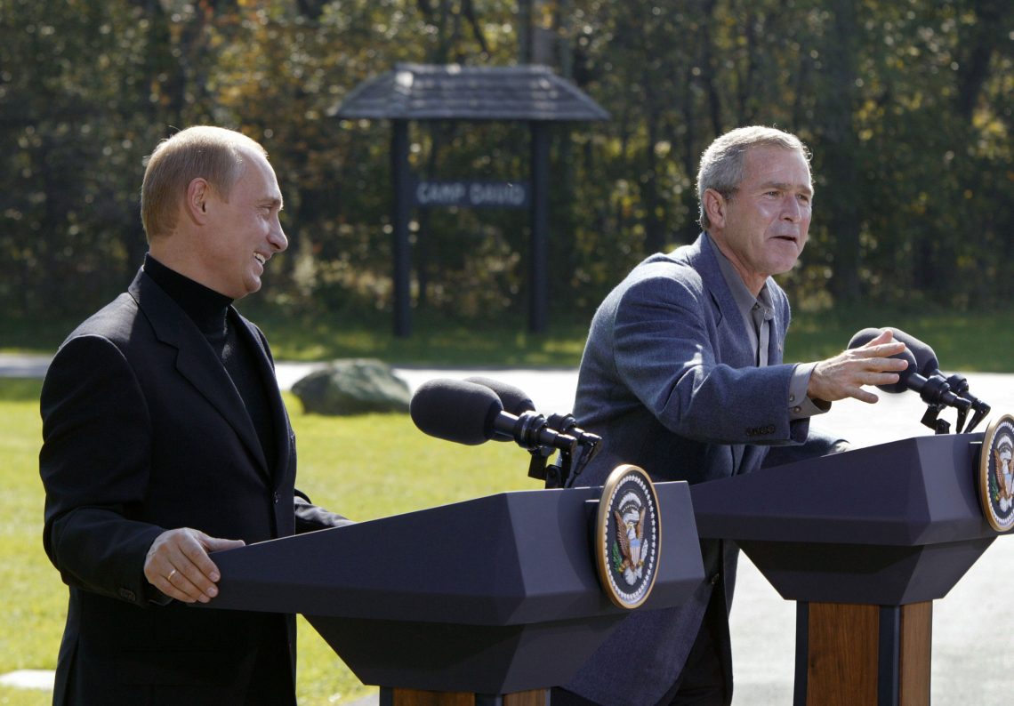Putin und George W. Bush stehen nebeneinander hinter zwei Rednerpulten mit dem Emblem der USA und lachen. Im Hintergrund ist eine überdachte Schautafel mit der Aufschrift Camp David zu sehen. Es geht um Atomwaffen und den Iran.