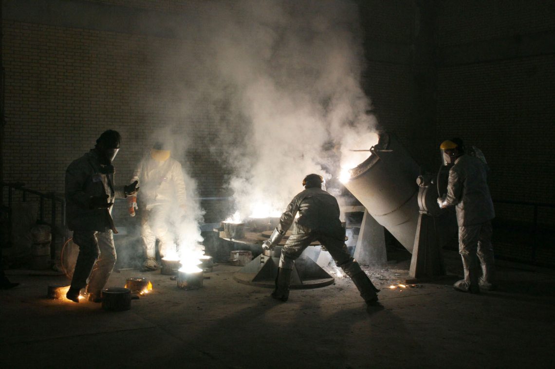 Im dichten Rauch im Gegenlicht stehen Männer in Schutzanzügen und hantieren mit industriellem Gerät. Das Bild illustriert einen Beitrag über Waffen im Iran.