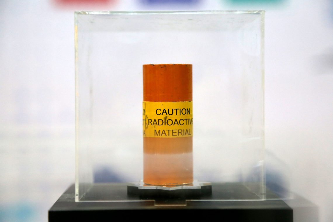 Ein orange-rot gestrifter zylindrischer Behälter in einem Plexiglas-Behältnis ist mit Caution Radioactive Material beschriftet.