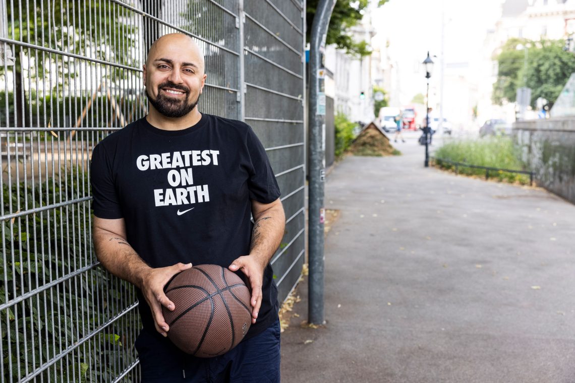 Foto von Ali Mahlodji, de rmit einem schwarzen T-Shirt mit der Aufschrift Greatest on Earth an einen Basketballkäfig gelehnt steht, einen Basketball in der Hand hält und in die Kamera lächelt. Das Bild illustriert ein Interview mit ihm .