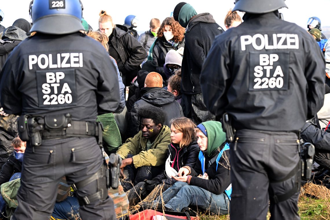 Foto von einer Gruppe Jugendlicher die von Polizisten umringt auf dem Boden sitzen. In der Mitte des Bildes GretaThunberg. Das Bild soll das Engagement von Jugendlichen für das Klima zeigen.