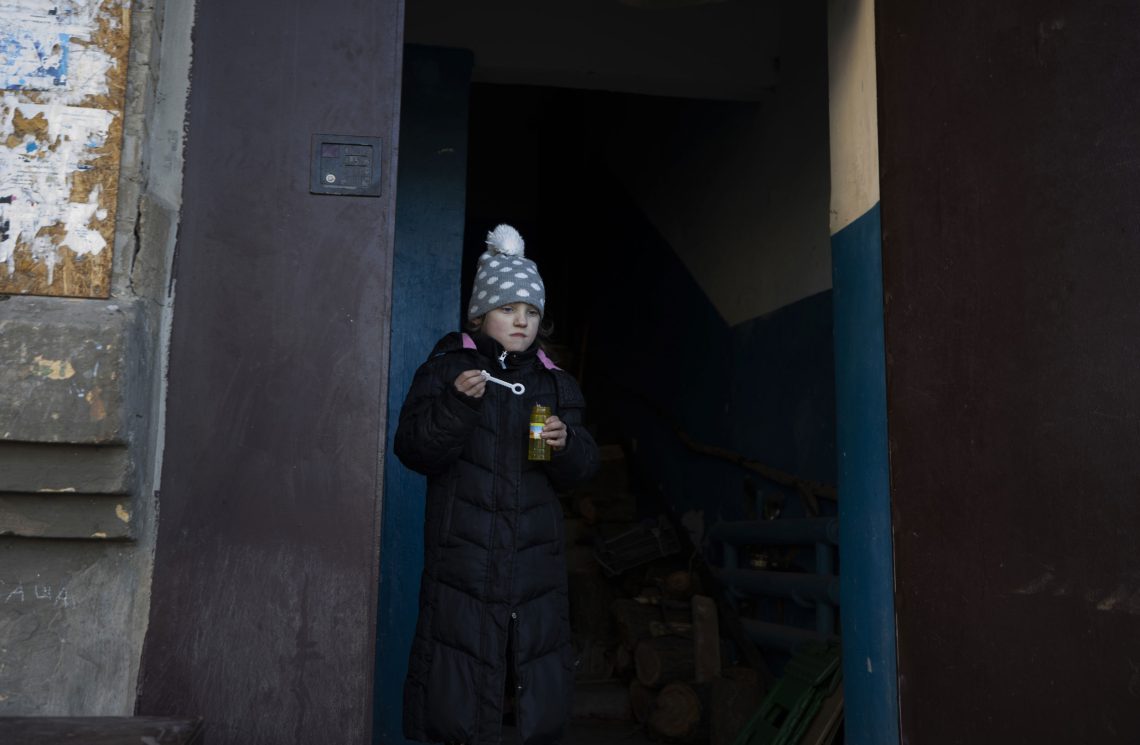Ein Mädchen mit einer grauen Mütze mit weißen Punkten steht vor einem Gebäude und ist dabei Luftblasen zu machen. Das Bild illustriert einen Beitrag über den Krieg in der Ukraine.