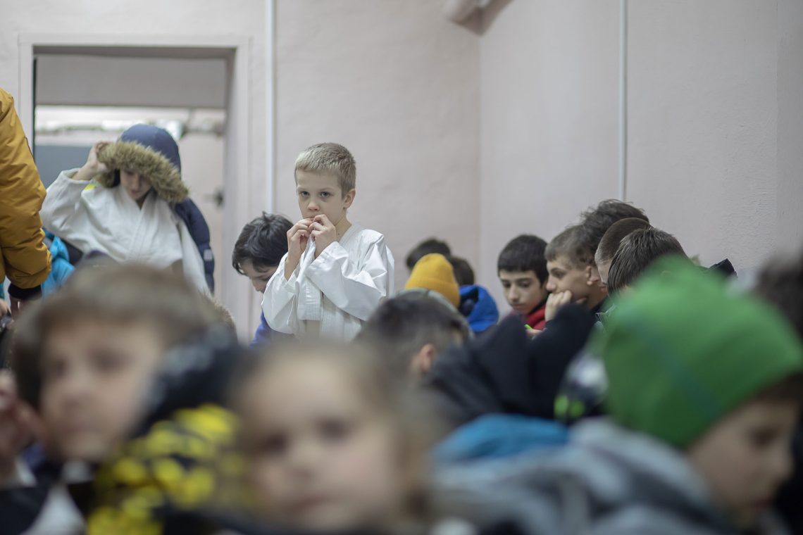 Eine Gruppe von Kindern unterschiedlichen Alters in einem weiß angestrichenen Raum. Ein Junge blickt in die Kamera, er trägt ebenso wie ein älteres Mädchen neben ihm einen Judoanzug. Das Bild zeigt ukrainische Schüler, die sich nach einem Luftangriff-Alarm in einem Schutzkeller verstecken. Es gehört zu einem Beitrag anlässlich von einem Jahr Ukraine-Krieg.