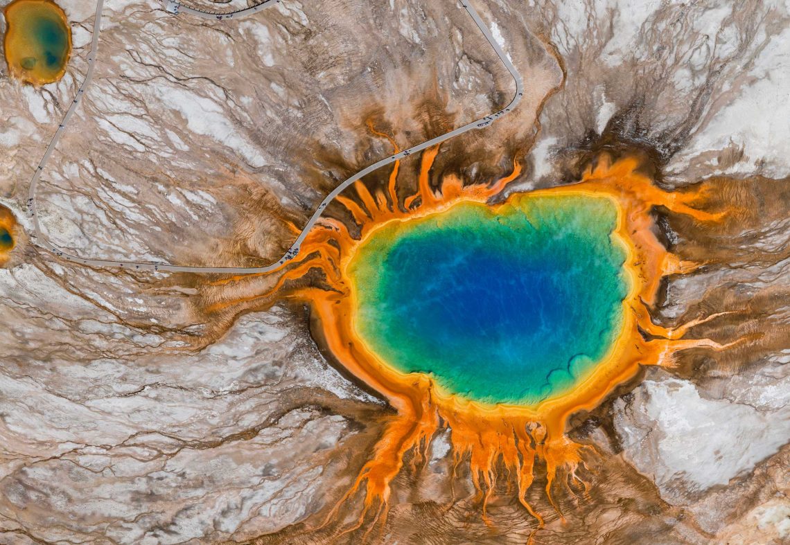 Foto aus der Vogelperspektive auf den fluoreszierend in blau, grün und orange leuchtenden See der Grand Prismatic Spring im Yellowstone Nationalpark. Die Farben stammen von Archaeen, die das heiße Wasser aushalten.