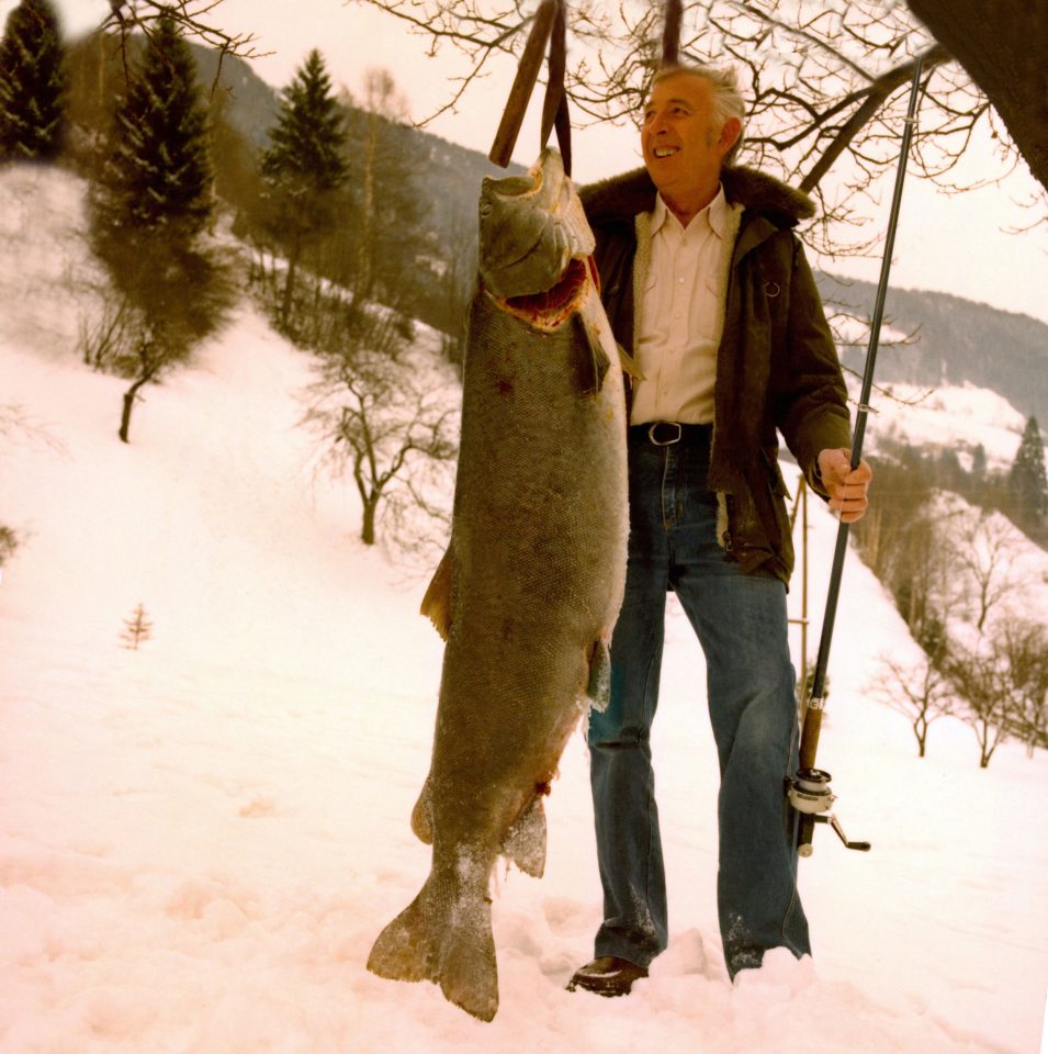 Ein Mann steht in einer Schneelandschaft und hält einen großen Fisch, der fast so groß ist, wie er selbst, mit der rechten Hand. In der Lingen eine Angel. Der Fisch ist ein Huchen. Eine ausgestorbene Fischart.