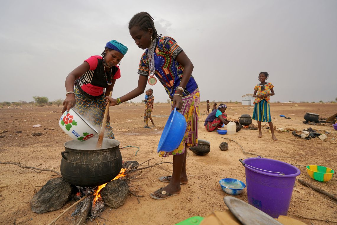 Foto von zwei Frauen, die bei offenem Feuer im Freien in einem großen Kessel eine Mahlzeit zubereiten. Im Hintergrund sind weitere Frauen und Kinder zu sehen. Das Bild ist Teil eines Beitrags über Burkina Faso.