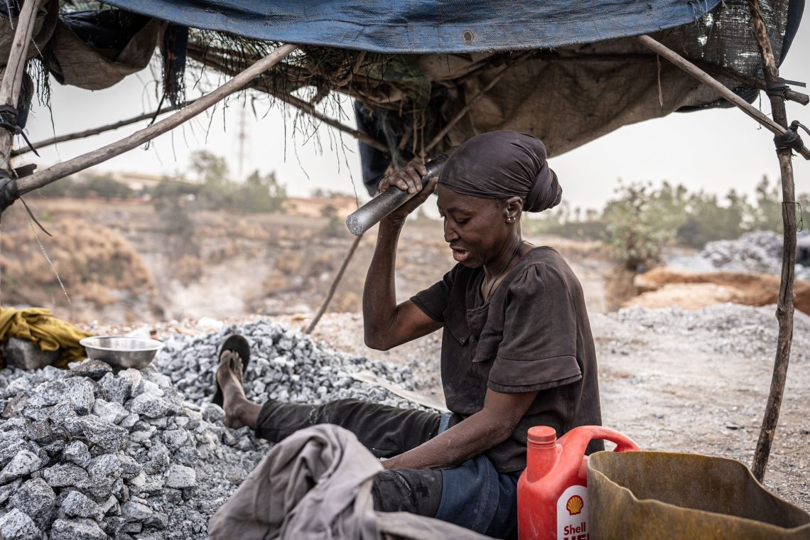 Ene Frau sitzt unter einer Stoffplane auf einem Steinhaufen und hält einen großen Stößel in der Hand mit der sie gerade ausholt, um Steine zu zerbrechen. Das Foto illustriert einen Beitrag über Burkina Faso, in dem es auch um Flucht geht.