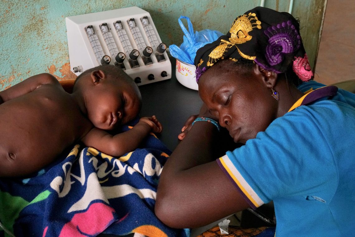 Eine Frau und ein kleines Kind schlafen, wobei die Frau mit dem Kopf auf den Armen abgestützt auf einem Tisch ruht. Das Kind liegt vor ihr und schläft. Es ist ein Foto, das in Burkina Faso aufgenommen wurde.