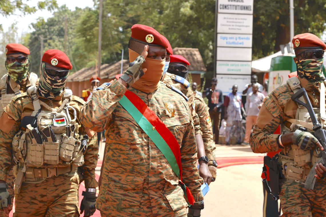 Ein salutierender Mann in Armee-Kleidung, das Gesicht halb bedeckt und mit rotem Beret salutiert in Begleitung von weiteren militärisch gekleideten Männern mit Waffen.