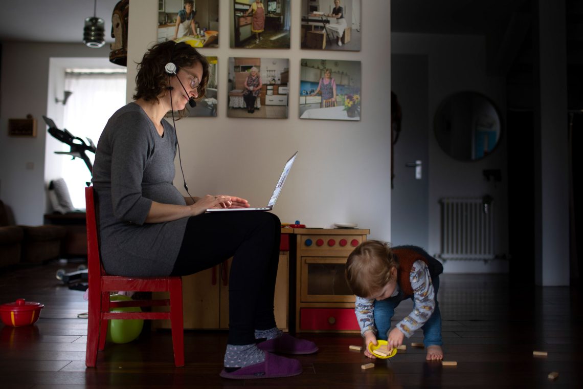 Eine Frau sitzt auf einem kleinen roten Schemel und tippt auf einem Laptop, während ein Kleinkind zu ihren Füßen spielt. Das Bild soll die aktuelle Aufteilung von Arbeit zwischen den Geschlechtern illustrieren und zeigen, dass Frauen in der Regel in den Familien mehr Arbeit leisten als Männer.