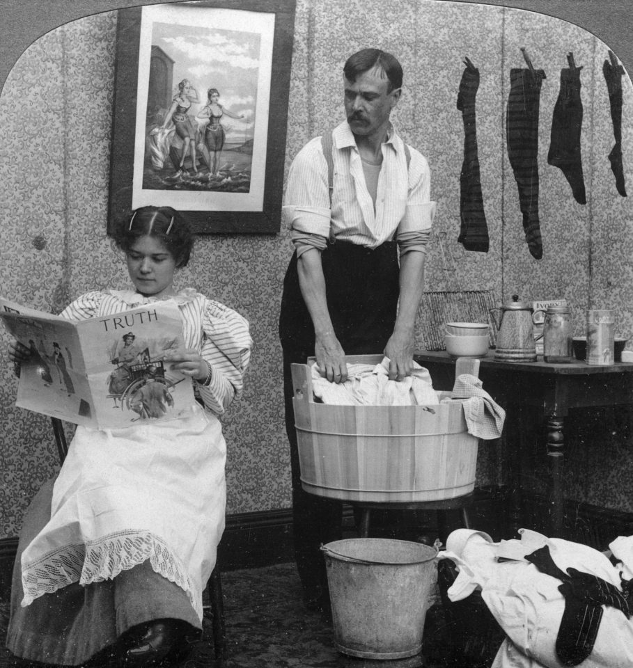 Eine Frau sitzt Zeitung lesend auf einem Stuhl während ein Mann an einem Waschzuber steht und Wäsche mit einem Waschbrett wäscht. Das Bild illustriert einen Kommentar von Christian Ortner, in dem es um Hausarbeit von Frauen in Familien geht.