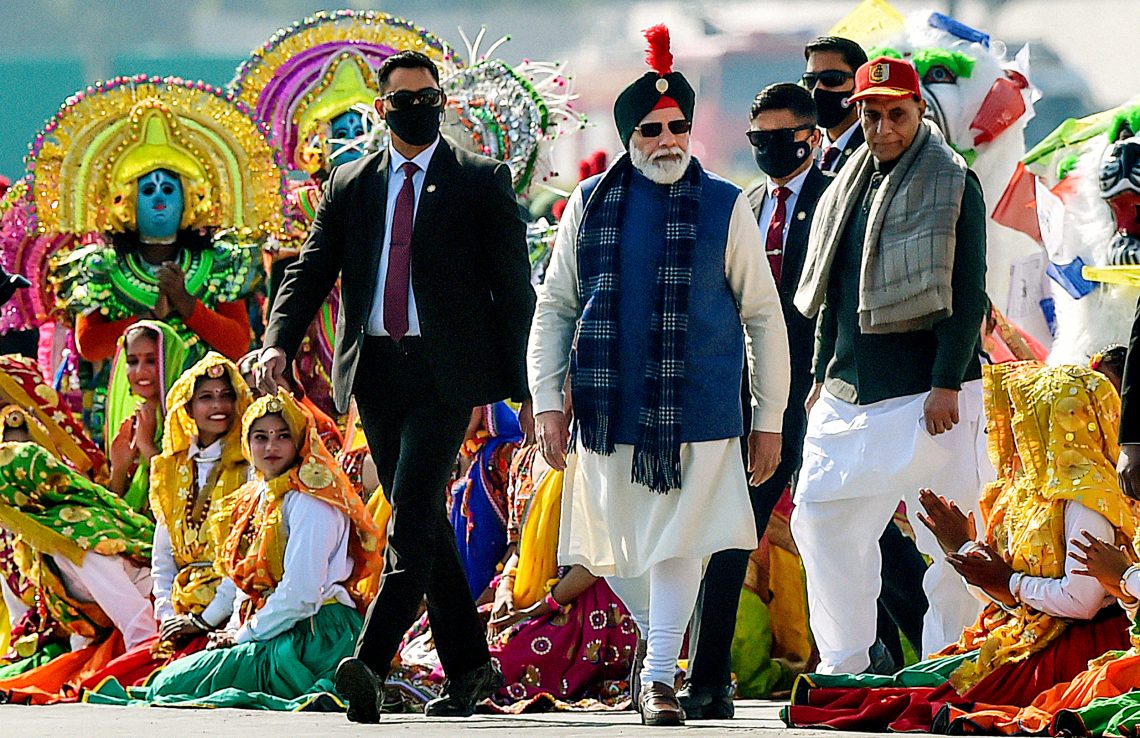 Indiens Premierminister Narendra Modi nimmt im Jänner 2022 in Neu-Delhi bei einer Feier von Militärkadetten anlässlich des 75-jährigen Jubiläums der nationalen Unabhängigkeit teil. Er trägt traditionelle Kleidung und Sonnenbrille, um ihn herum sitzen Frauen, gekleidet in den Nationalfarben und im Hintergrund sind als Gottheit Krishna kostümierte Gestalten.
