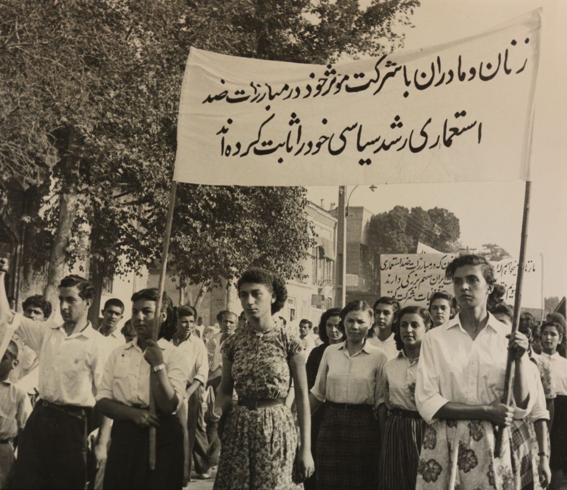 Schwarz-weiß Fotografie von Frauen und jungen Männern die bei einer Demonstration ein großes Banner mit persischen Schriftzügen tragen und ernst blicken.