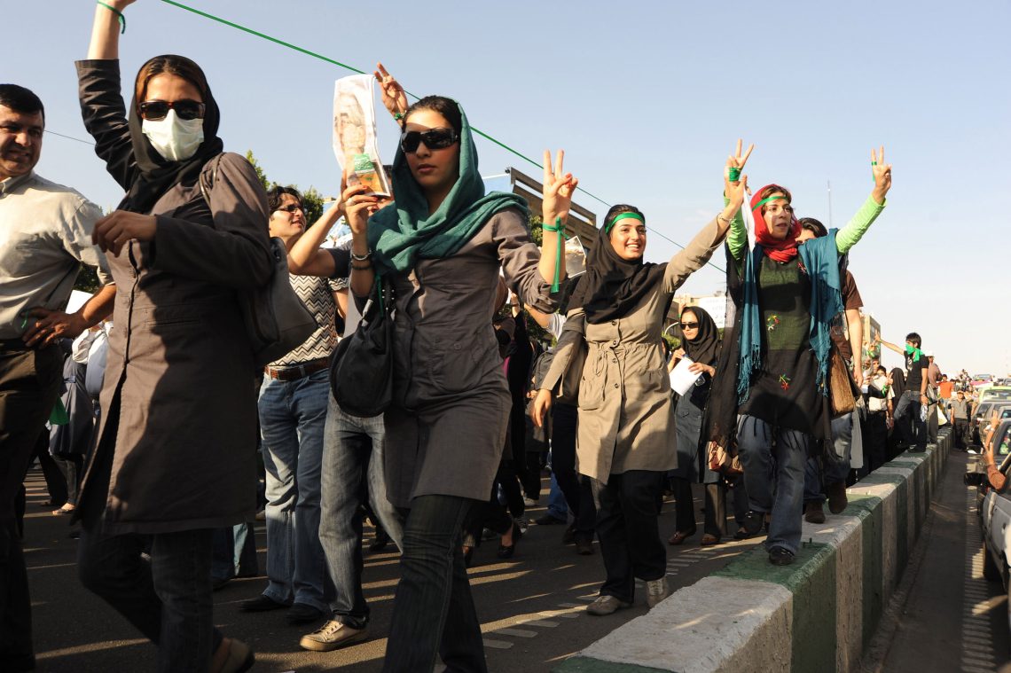Frauen mit zum Victoryzeichen erhobenen Händen gehen eine Straße entlang und halten Fotos von Mir Hossein Mousavi, einem Präsidentschaftskanidaten, in die Höhe.