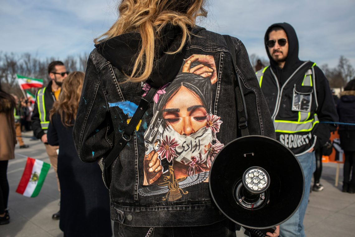 Foto einer Frau mit einer schwarzen Jeansjacke, auf deren Rücken ein Bild von einer Frau mit geschlossenen Augen und einer Mundbedeckung abgebildet ist Die Frau hält ihre Haare fest. Die Geste zeigt, dass die Frau sich die Haare abschneiden will, ein Symbol für die Revolution der Frauen im Iran 2023.