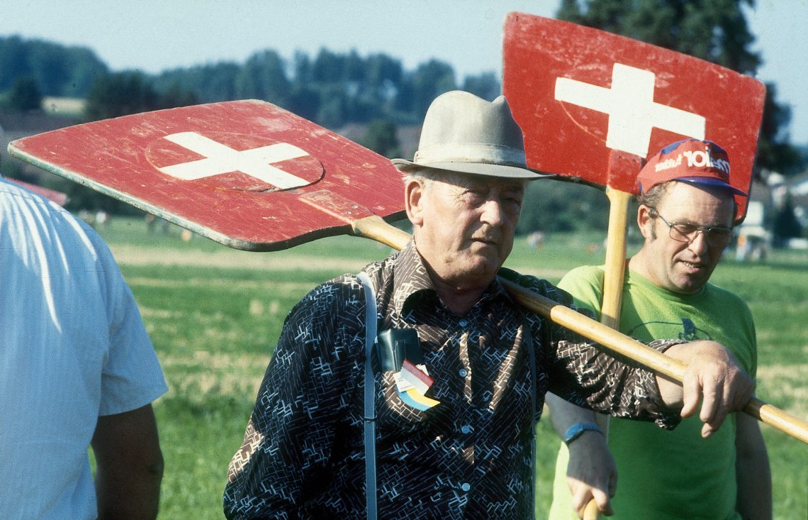 Historische Fotografie von Schweizer Bauern beim Hornussen auf einem Feld 1979. Sie halten große Holzschaufeln mit dem Schweizerkreuz darauf. Damit müssen sie ein Geschoss, das die Gegenmannschaft mit einem flexiblen Schläger über ihre Köpfe hinweg peitscht, abfangen..