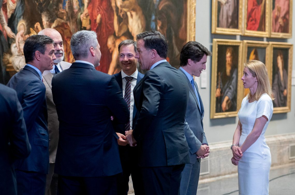 Foto einer Gruppe von Männern in Anzügen und einer Frau, die am Rand steht und ein weißes Kleid trägt. Ein Mann hat der Männergruppe den Rücken zugekehrt und spricht mit der Frau. Diese ist die Premierministerin von Estland, Kaja Kallas. Das Bild ist Teil eines Beitrags über feministische Außenpolitik.