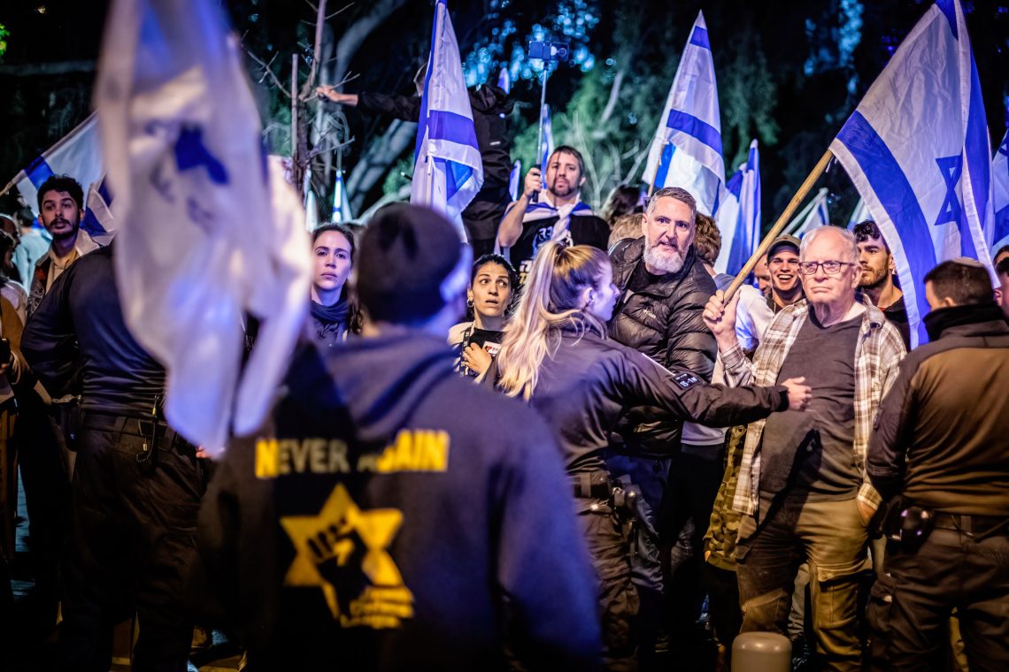 Foto einer Straßendemonstration in der abendlichen Dunkelheit, wobei eine Polizistin zwei Gruppen von einenr trennt. Die Proteste richten sich gegen die Justizreform in Israel. Die Gegner befürchten, dass die Demokratie in Israel durch die Reform beendet wird.