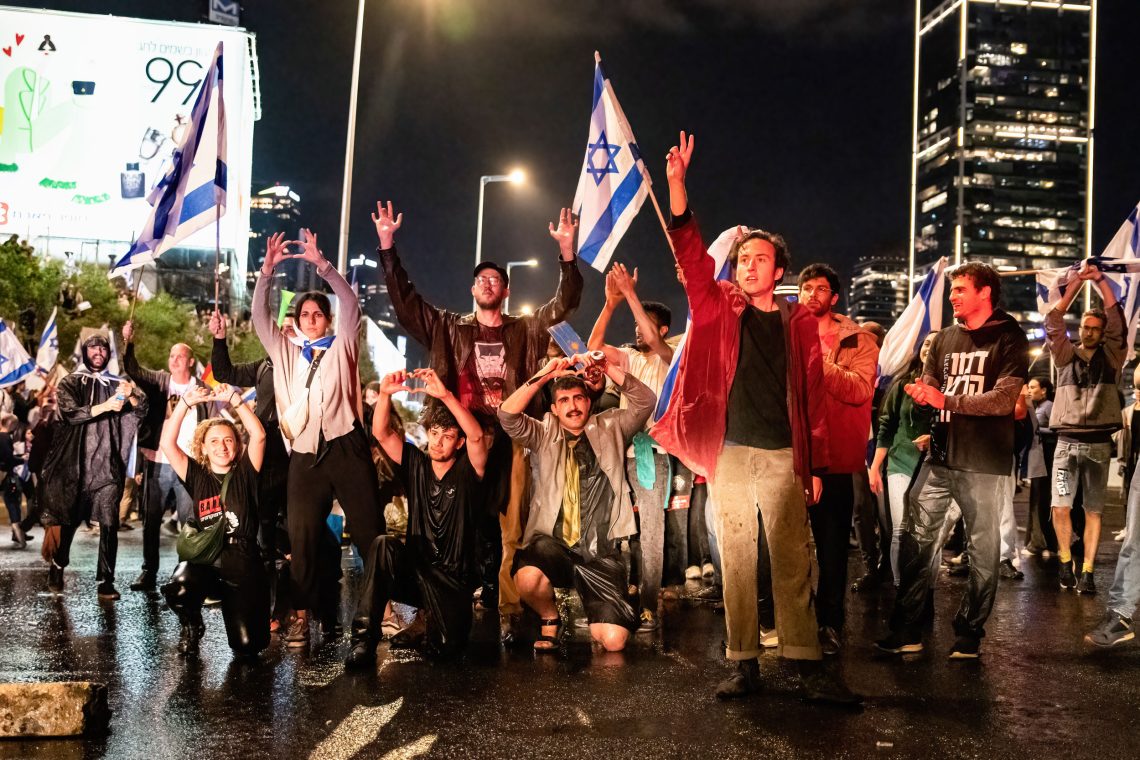Eine Gruppe von Protestierenden schwenkt Fahnen und formt mit den Händen Herzen. Es ist eine Demonstration für Demokratie in Israel.