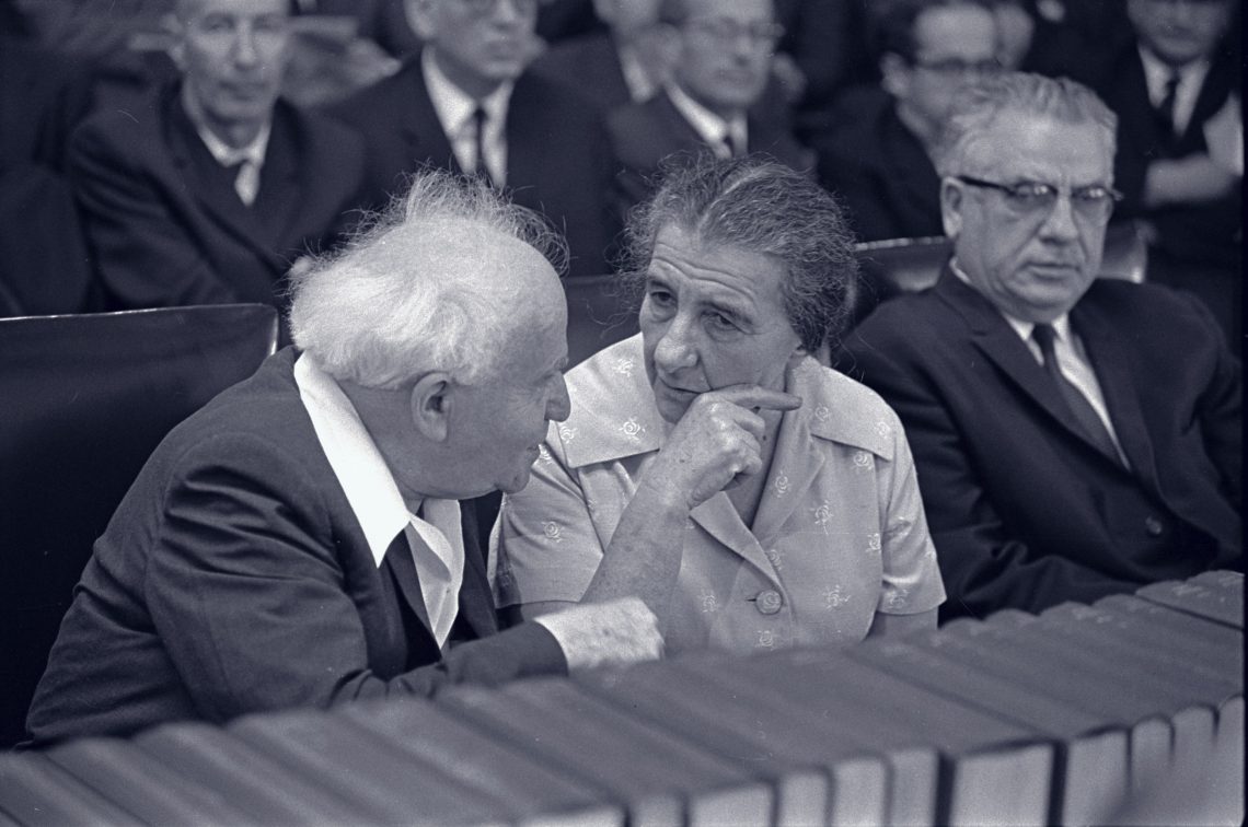 Schwarzweiß Foto von David Ben-Gurion und Golda Meir, die nebeneinander sitzen und diskutieren. Im Vordergrund stehen Bücher.