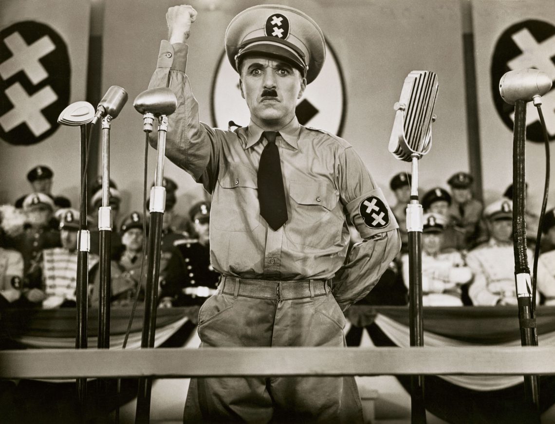 Charlie Chaplin in Der große Diktator als Adenoid Hynkel bei einer Rede. Neben ihm Mikrophone, im Hintergrund Fahnen mit zwei X statt dem Hakenkreuzsymbol. Der Film ist ein Beispiel für Witze über Diktatoren.