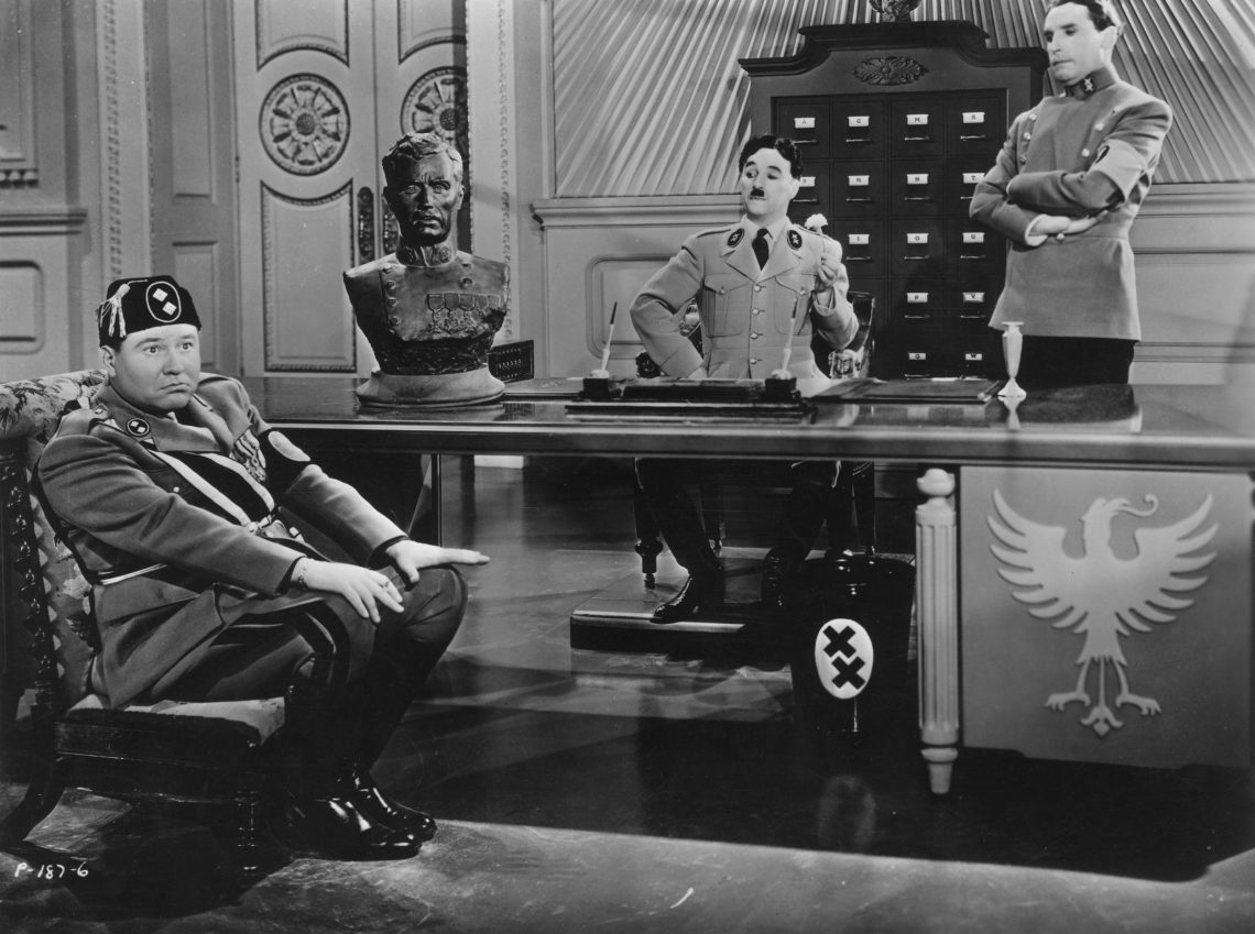 In der Szene aus „Der große Diktator“ sitzt Benito Mussolini auf einem so niedrigen Stuhll, dass er kaum über den Tisch blicken kann, hinter dem Charlie Chaplin als Diktator Adenoid Hynkel auf einem erhöhten Stuhl sitzt und auf ihn herabblickt.