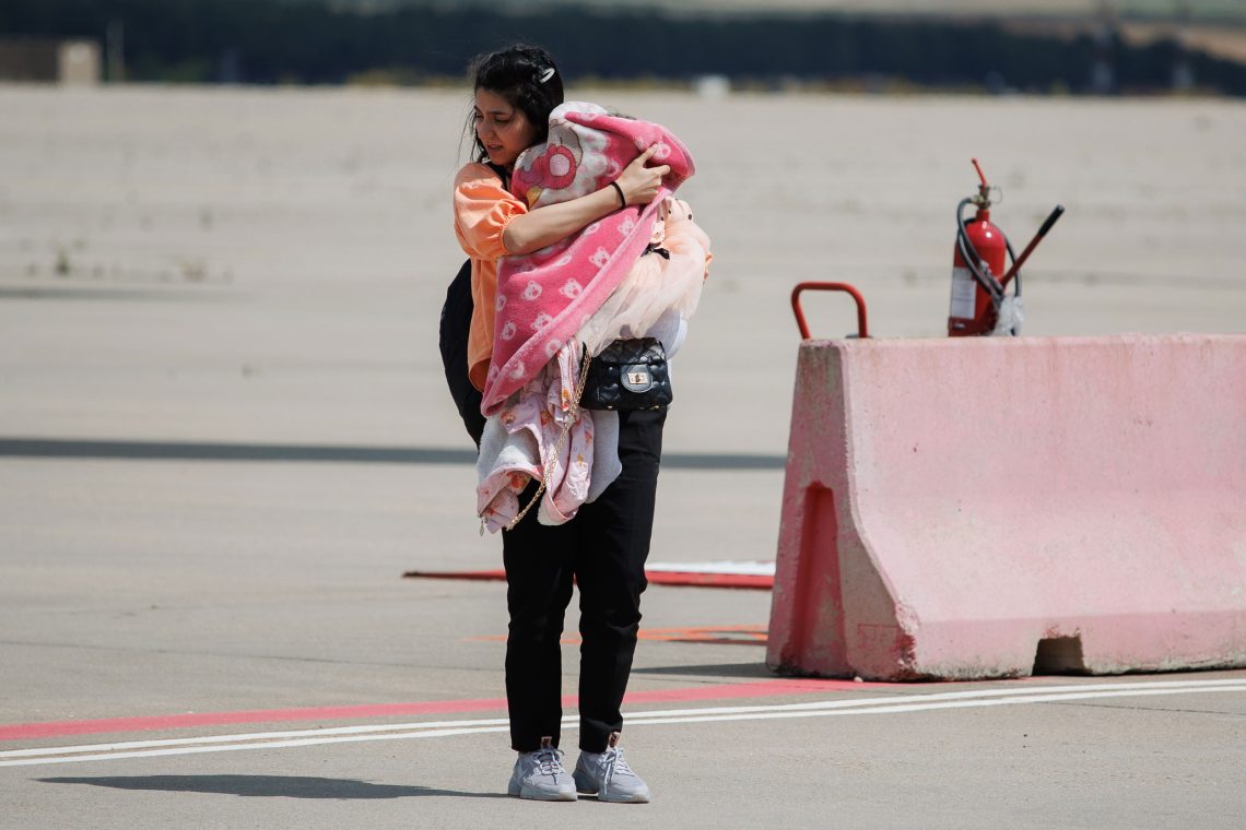 EIne Frau in Turnschuhen und heller Bluse hält ein Baby in Decken gewickelt in den Armen und blickt zur Seite auf den Boden. Das Bild illustriert einen Beitrag über Flucht und Asylrecht.