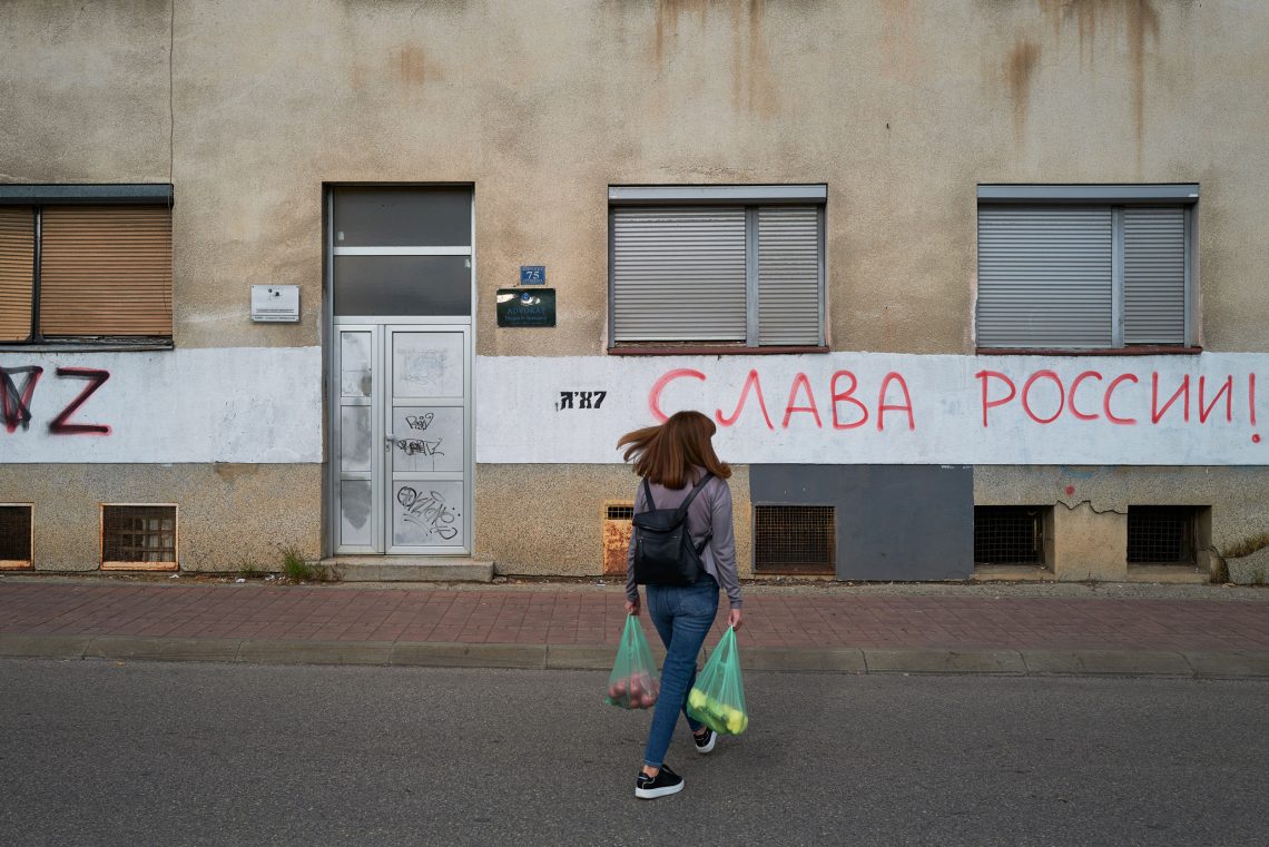 Eine Frau mit langen Haaren überquert zwei Einkaufstaschen tragend eine Straße. Das Bild illustriert einen Beitrag über Russland und den Balkan.