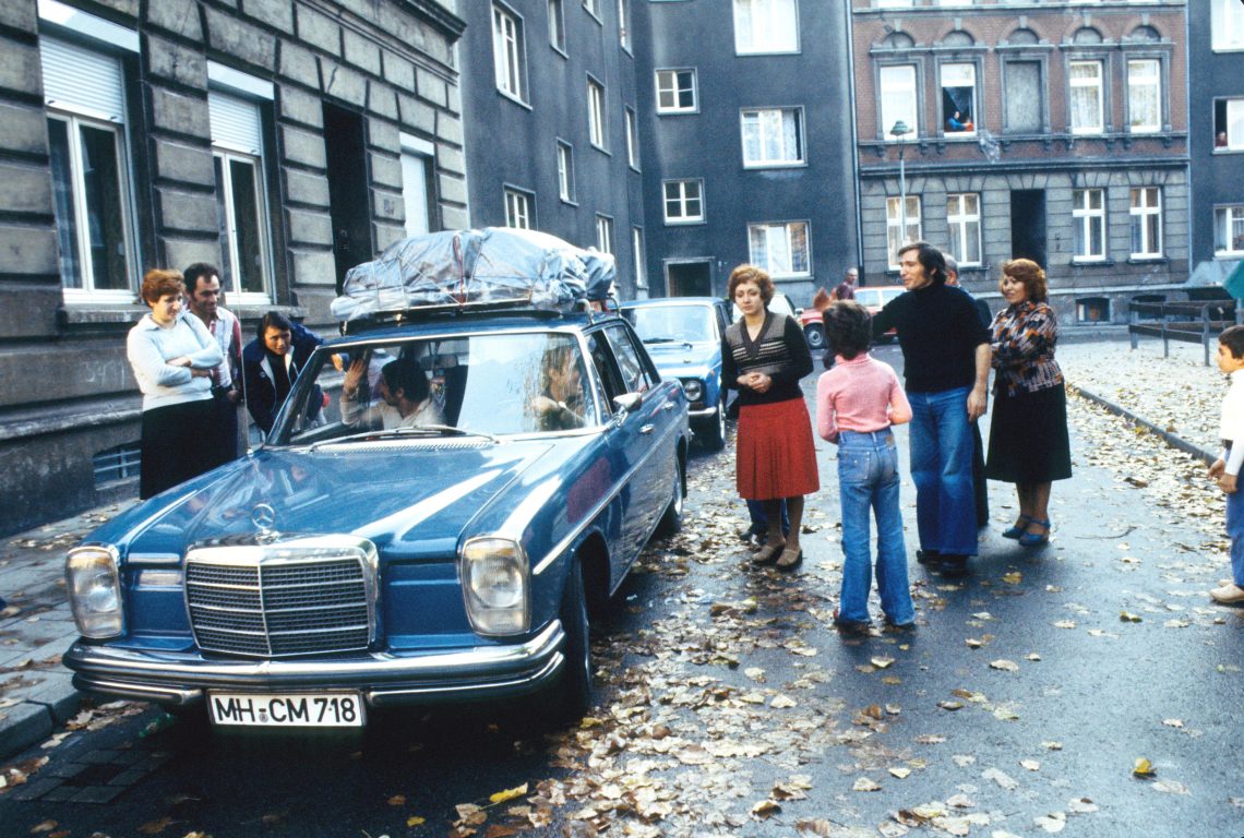 Ein blauer Mercedes mit Gepäck auf dem Dach steht auf einer Straße in einem städtischen Wohngebiet umringt von einer Gruppe Menschen, die sich zum Fahrer im Auto herunterbeugen. Das Bild illustriert einen Beitrag über Zuwanderung und Handwerk