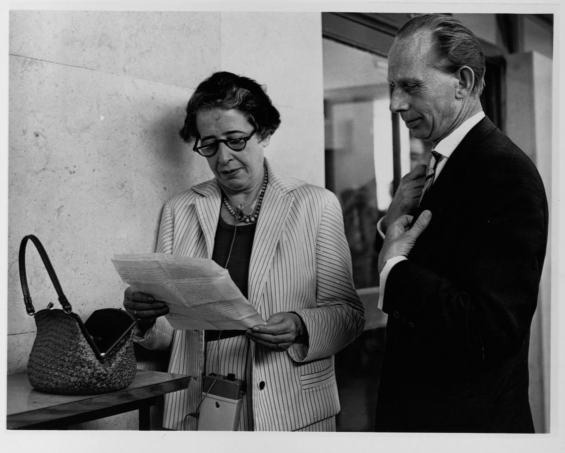 Hannah Arendt in einem hellen Kostüm steht vor einem Tisch und liest ein Dokument. Neben ihr steht ein Mann. Die Szene scheint vor einem Vortrag stattzufinden. Das Bild illustriert einen Beitrag über die Verfahren in einem Asylantrag.