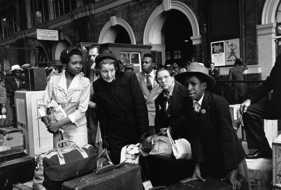 Eine Menschengruppe mit Koffern und Taschen in einem großen Gebäude, eine Frau in der Mitte trägt den für die Heilsarmee typischen Hut.