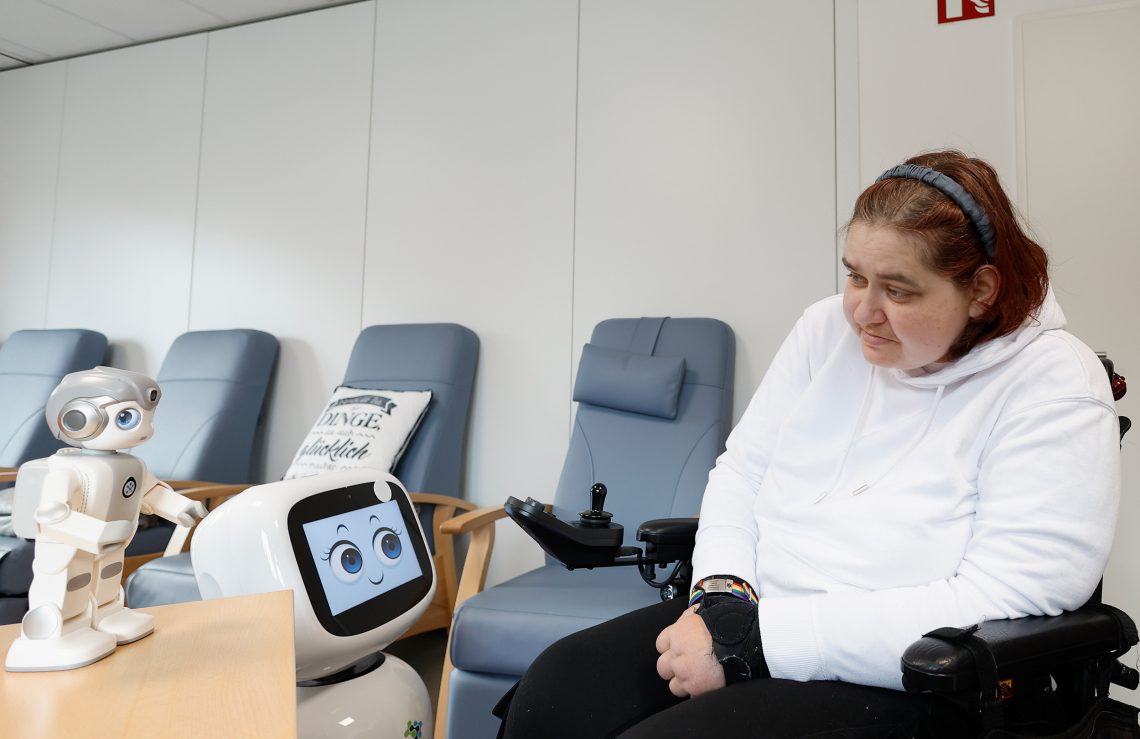 Ein Roboter mit einem Display-Gesicht hält Blickkontakt mit einer Patientin im Rollstuhl in einem Pflegeheim.