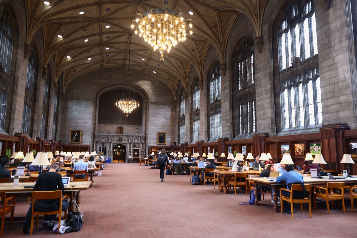 Lesesaal der Bibliothek der Universität Chicago mit rotem Teppisch und einer neugothischen Halle mit großen Fenstern und riesigen Kronleuchtern sowie Tischen mit Leselampen. Die Menschen sitzen an Laptops und Büchern.