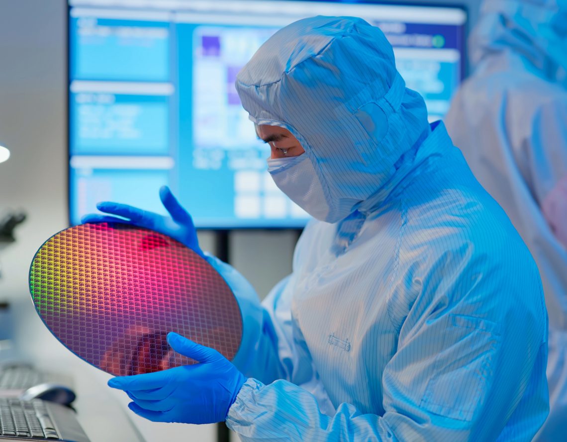 Ein Techniker in einem sterilen Ganzkörperanzug wirft einen prüfenden Blick auf eine bunt schillernde Halbleiterplatte.