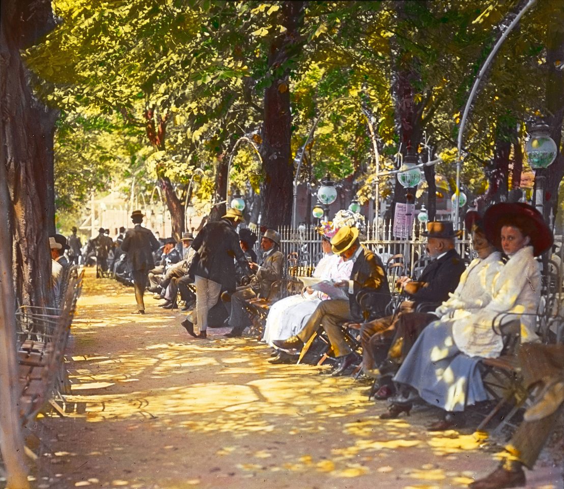 nachträglich kolorierte Aufnahme von Menschen, die unter großen Bäumen sitzen und Zeitung lesen. Die Männer tragen Strohhüte.