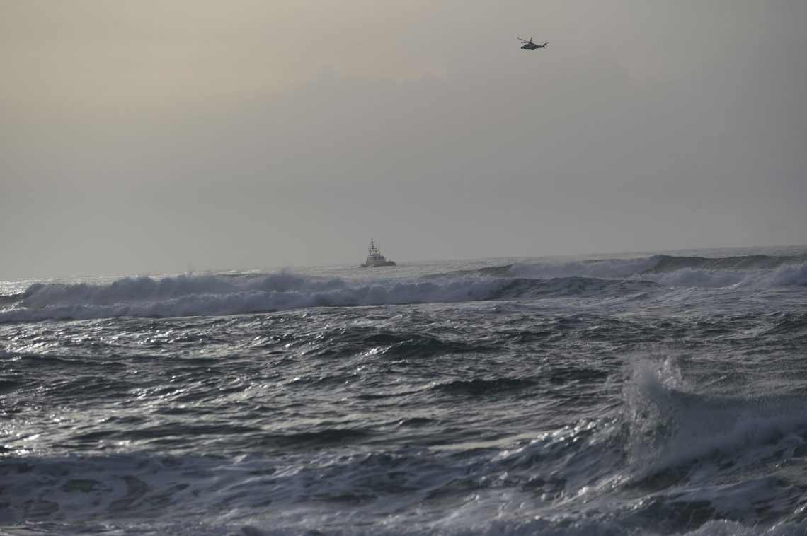Das aufgewühlteMittelmeer vor der italienischen Küste mit Hubschrauber und Boot. Das Bild ist Teil eines Kommentars zum Thema Rückführung bzw. Abschiebung von Asylsuchenden.