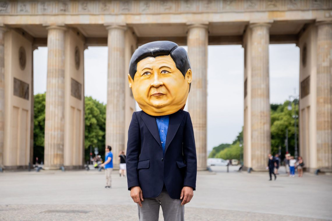 Ein Mann mit blauer Anzugjacke trägt einen großen Kopf aus Pappmaché, der den chinesischen Staatspräsidenten Xi Jinpung darstellt, auf dem Kopf. Im Hintergrund ist das Brandenburger Tor zu sehen. Das Bild illustriert einen Beitrag über die Machtpolitik Chinas und die Seidenstraße bzw. die Neue Seidenstraße.