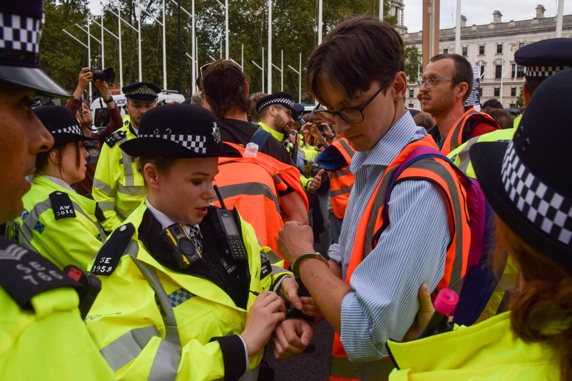 Verhaftung eines Mannes mit Brille udn gestreiftem Hemd durch eine Polizistin, die ihm Handschellen anlegt. Das Bild illustriert einen Beitrag üer Scheindemokratie bzw. die Bedrohungen, denen Demokratien heute ausgesetzt sind.