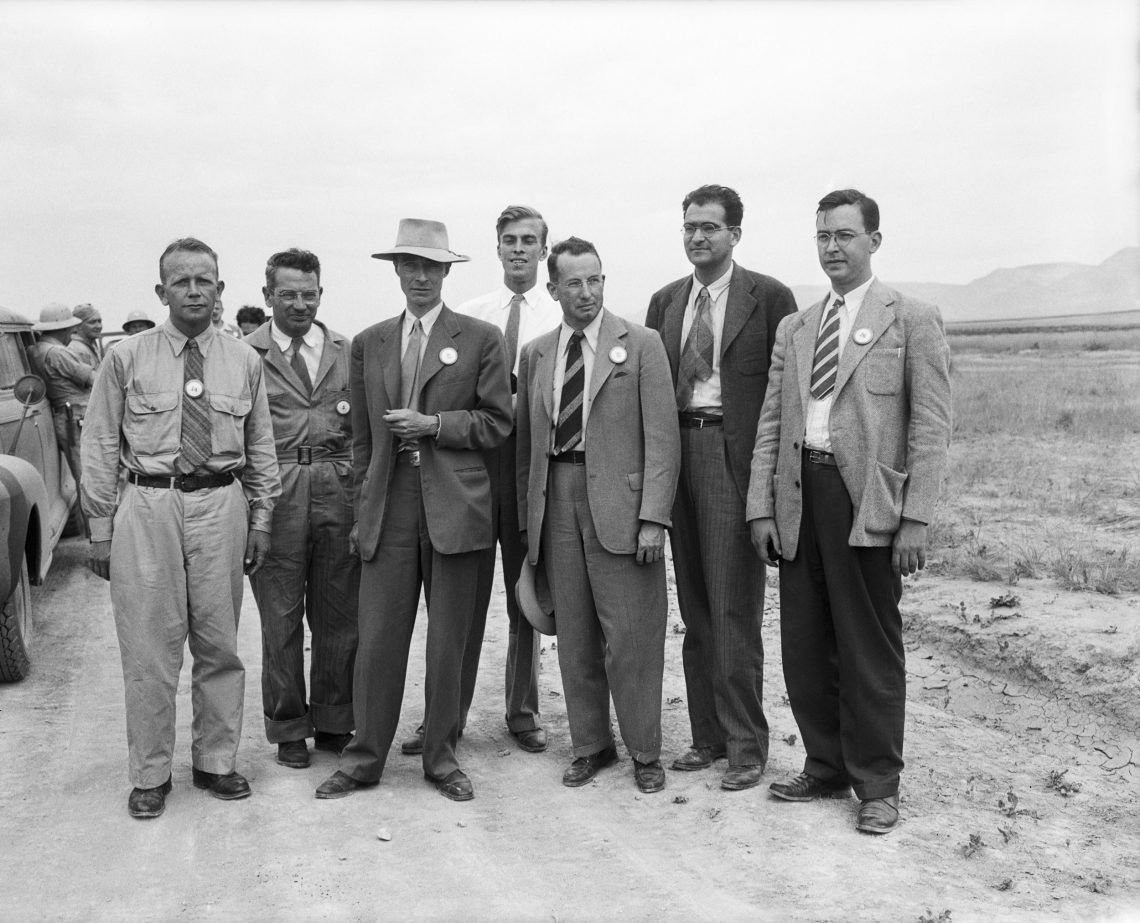 Sieben Männer in Anzügen bzw. Uniformähnlichen Outfits posieren in einer steppenartigen Landschaft. In der Mitte steht der Physiker Robert Oppenheimer mit Hut. Das Bild ist Teil eins Beitrags über die Frage, ob der Abwurf der Atombomben ein Kriegsverbrechen war.