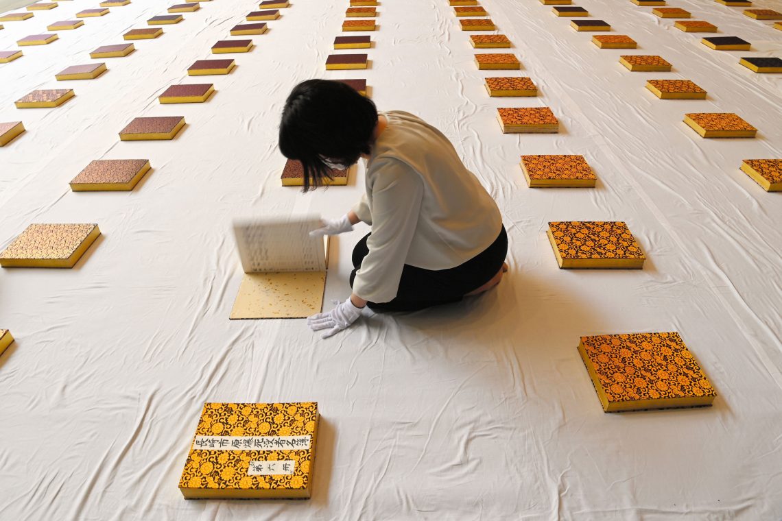 Eine Frau sitzt auf einem weißen Tuch umgeben von Büchern mit einem Ornament, die in Reihen nebeneinander liegen. Die Frau trägt Handschuhe und hat eines der Bücher aufgeschlagen. Es handelt sich um eine Gedenkstätte an die Todesopfer des Atombombenabwurfs in Nagasaki. Die Bücher sind sogenannte Namensbücher.
