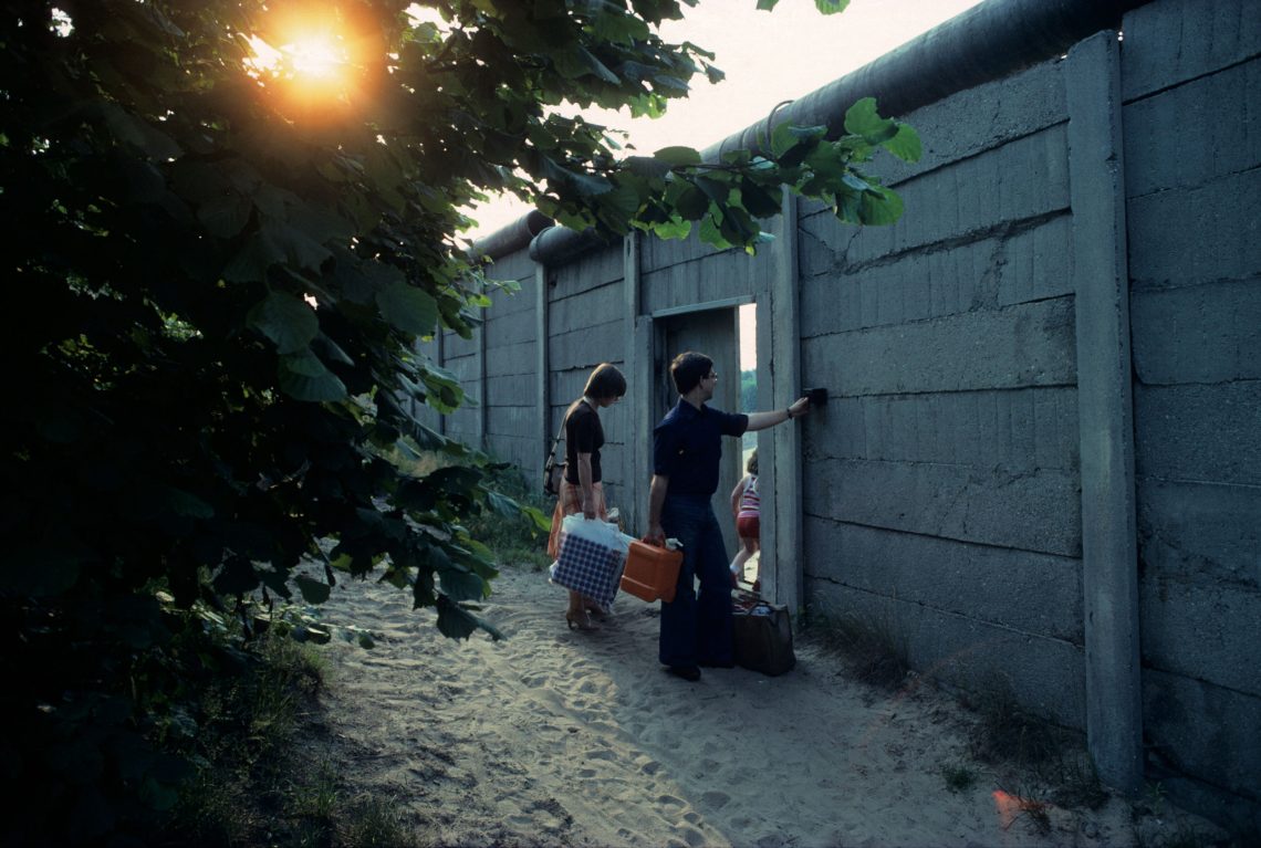 Ein Foto der Berliner Mauer in einem ländlichen Teil Berlins. Eine Familie mit Kind geht durch ein Tor in der Mauer. Dahinter ist viel Grün zu sehen, eine Schrebergartensiedlung.