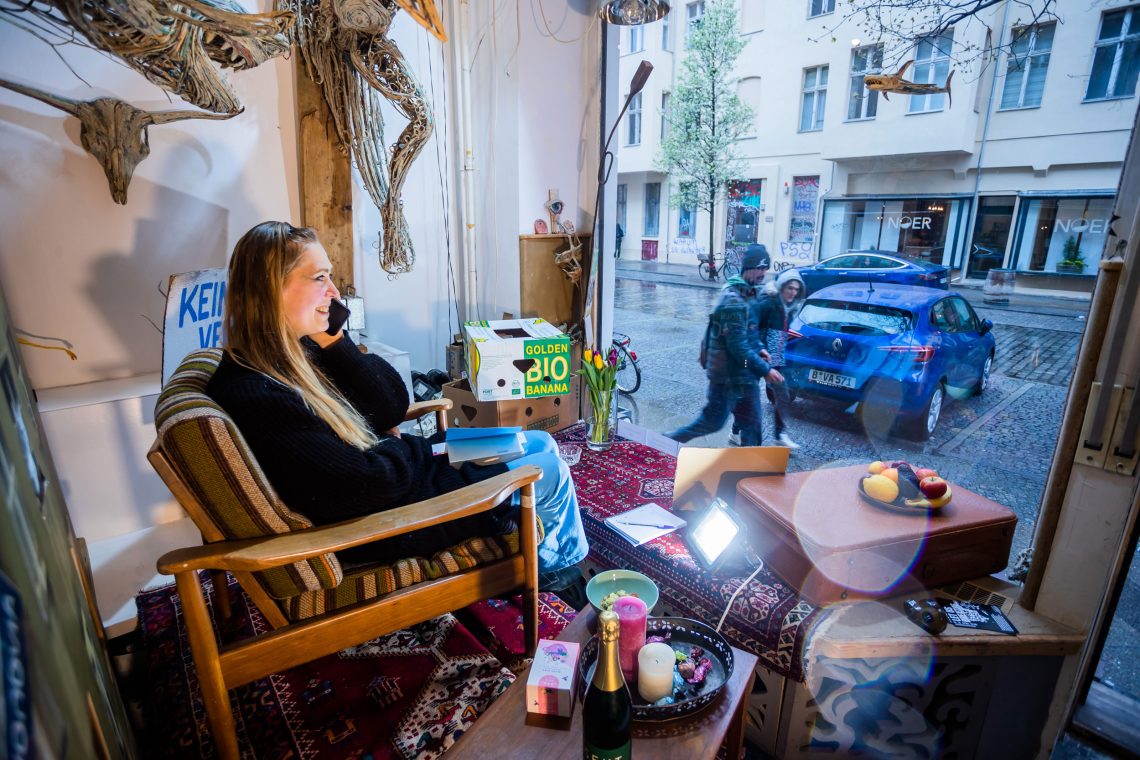Eine Frau sitzt telefonierend in einem Sessel. Die Künstlerlin hat sich gemütlich im Schaufenster eingerichtet, vor dem Menschen vorbeigehen. Die Sehnsucht nach dem Landleben hat oft Gründe in den hohen Wohnungskosten in den Städten.