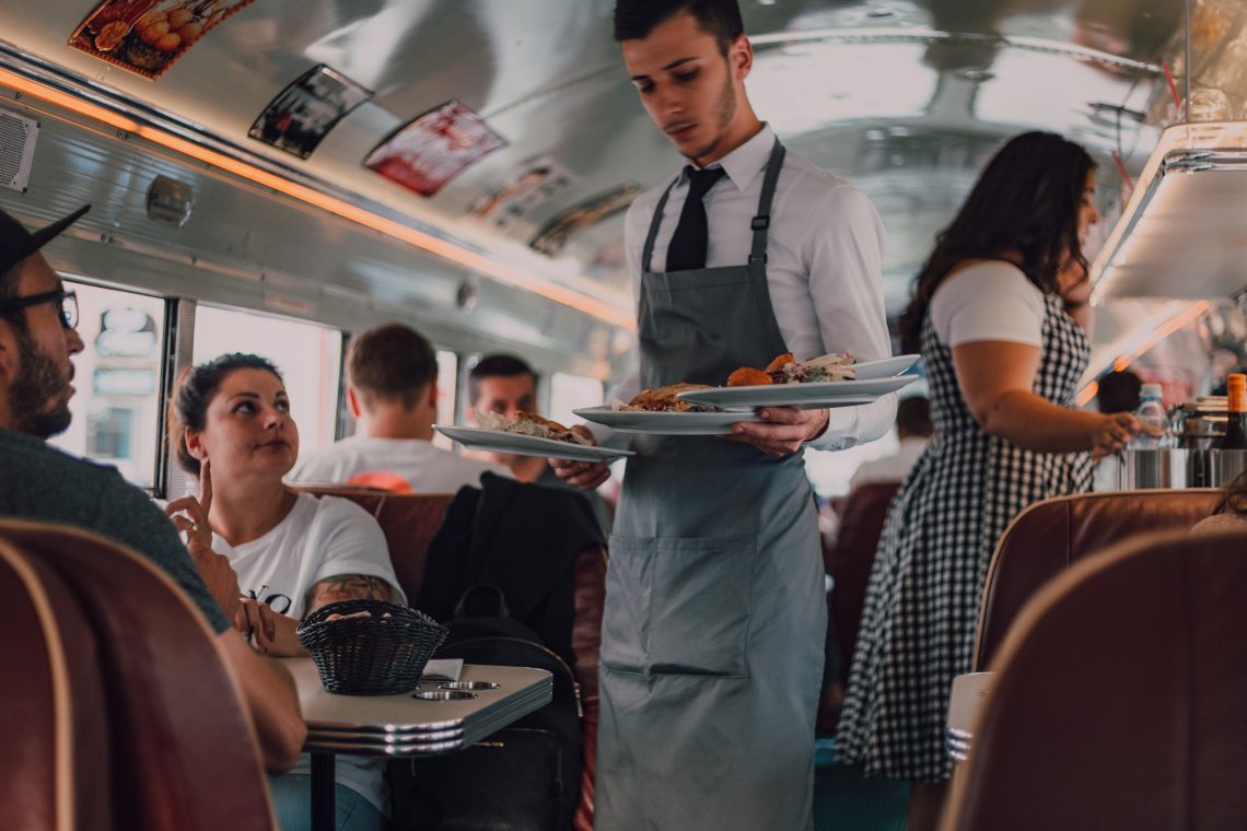 Ein Kellner mit weißen Hemd, Krawatte und grauer Schürze trägt vier reich beladene Teller an einen kleinen Tisch zu den Gästen. All das geschieht auf engstem Raum in einem umfunktionierten und fahrenden Schulbus.